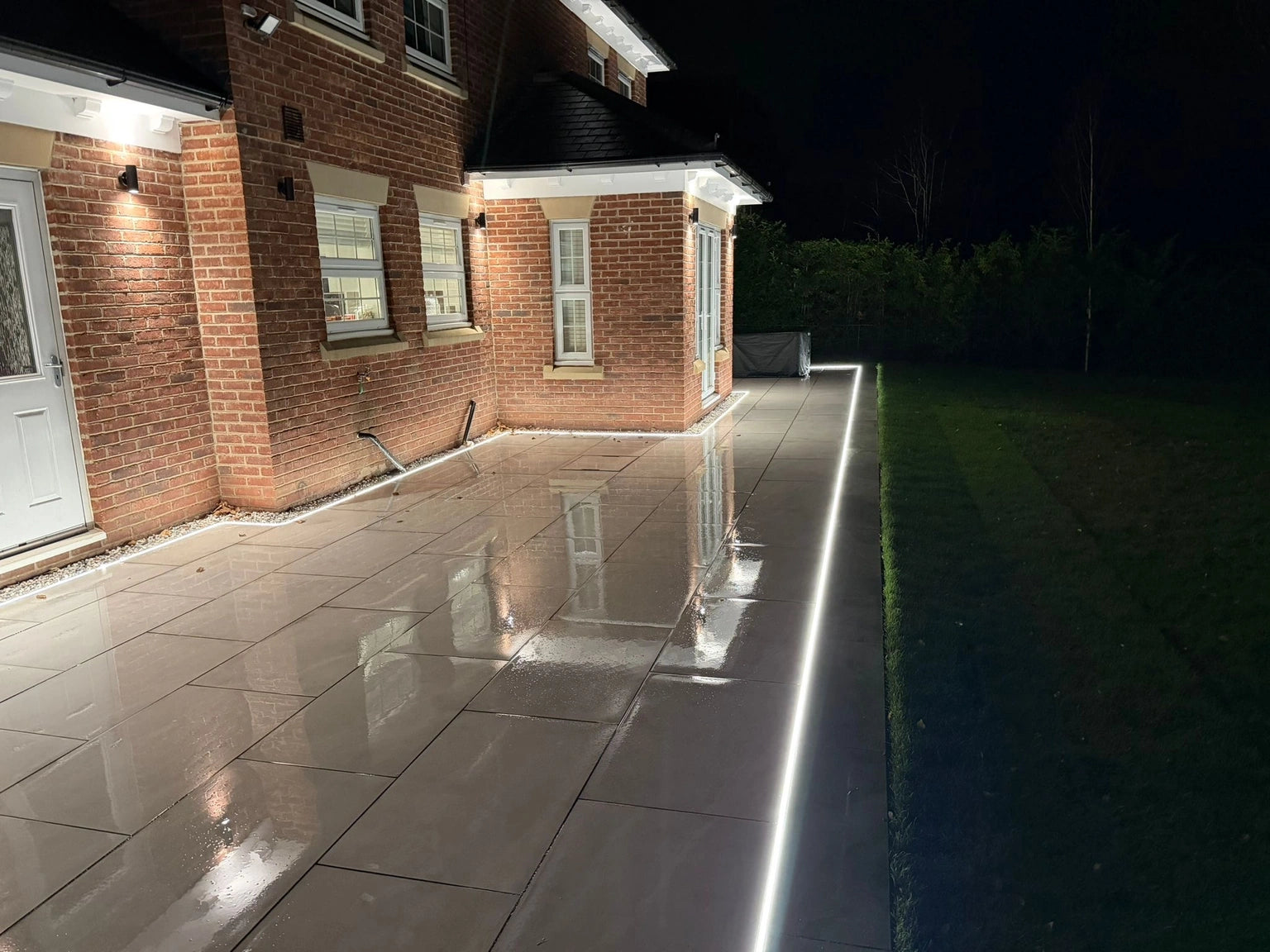 Recessed neon flex tracking down the patio at the property edge, wet paving reflecting the light